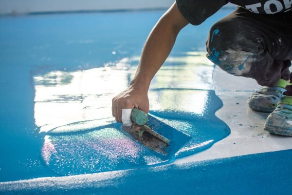 A person using a paint brush to apply blue color to a floor, highlighting epoxy flooring service in Austin, TX.